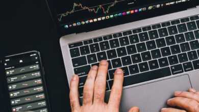 Hands typing on a laptop with cryptocurrency charts and smartphone display in view, focusing on digital finance.