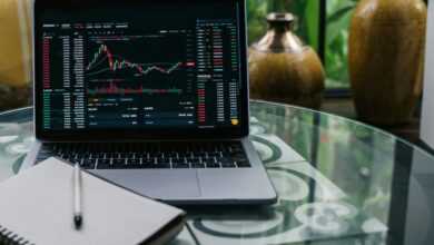 Laptop displaying cryptocurrency stocks and graphs on a glass table with a notepad.